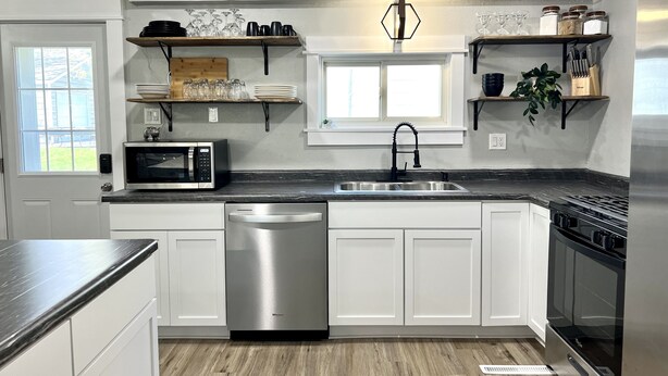 A modern kitchen with white cabinets, a stainless steel dishwasher, a microwave, a sink, and a stove. There are shelves above the counter holding dishes and glasses.