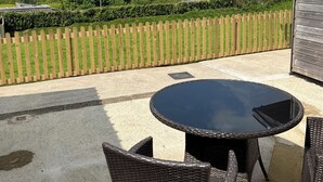 A patio with wicker furniture and a glass table, overlooking a green landscape with a wooden fence and a building in the background. - Coastal retreat Converted Stables, ,  close to Maidencombe beach & pubs. (Stokeinteignhead Torquay,)