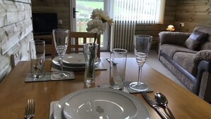 A dining table set with plates, glasses, and cutlery, a vase with flowers, and a view of a wooden cabin and greenery. - Coastal retreat Converted Stables, ,  close to Maidencombe beach & pubs. (Stokeinteignhead Torquay,)