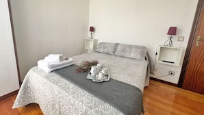 Ein ordentlich bezogenes Bett mit einer grau gemusterten Tagesdecke, ein Tablett mit Tassen und eine Blumenkomposition sowie ein weißer Nachttisch mit Lampe.