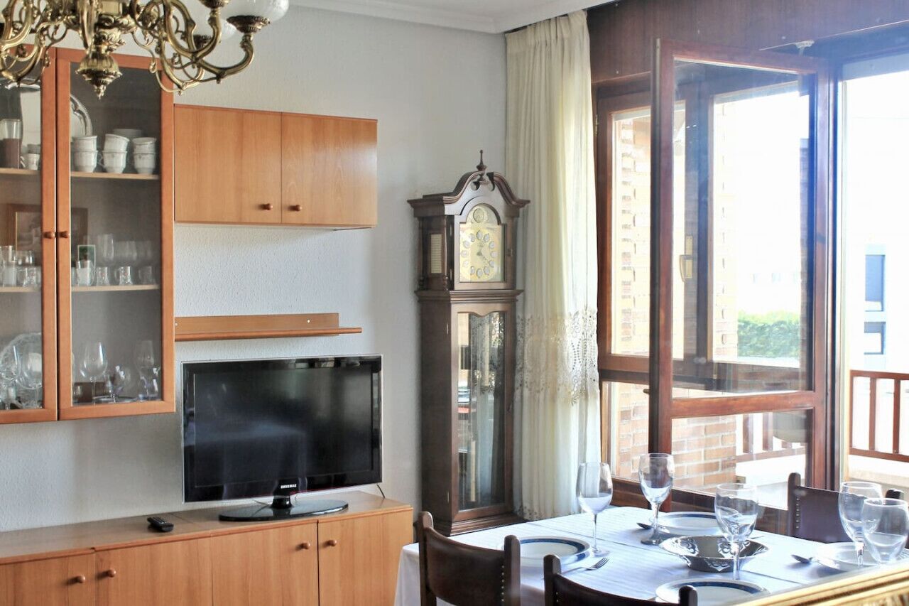 Una habitación con televisión, armarios de madera, un reloj y una mesa de comedor con vasos.