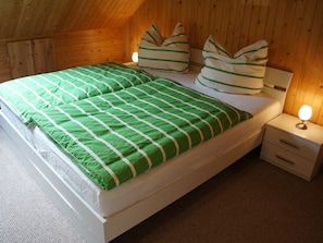 Ein Zimmer mit einem Einzelbett, geneigter Decke, Holzwänden, einer grün-weiß gestreiften Bettdecke, zwei Kissen, einem Nachttisch mit Lampe und einer kleinen Schublade.
