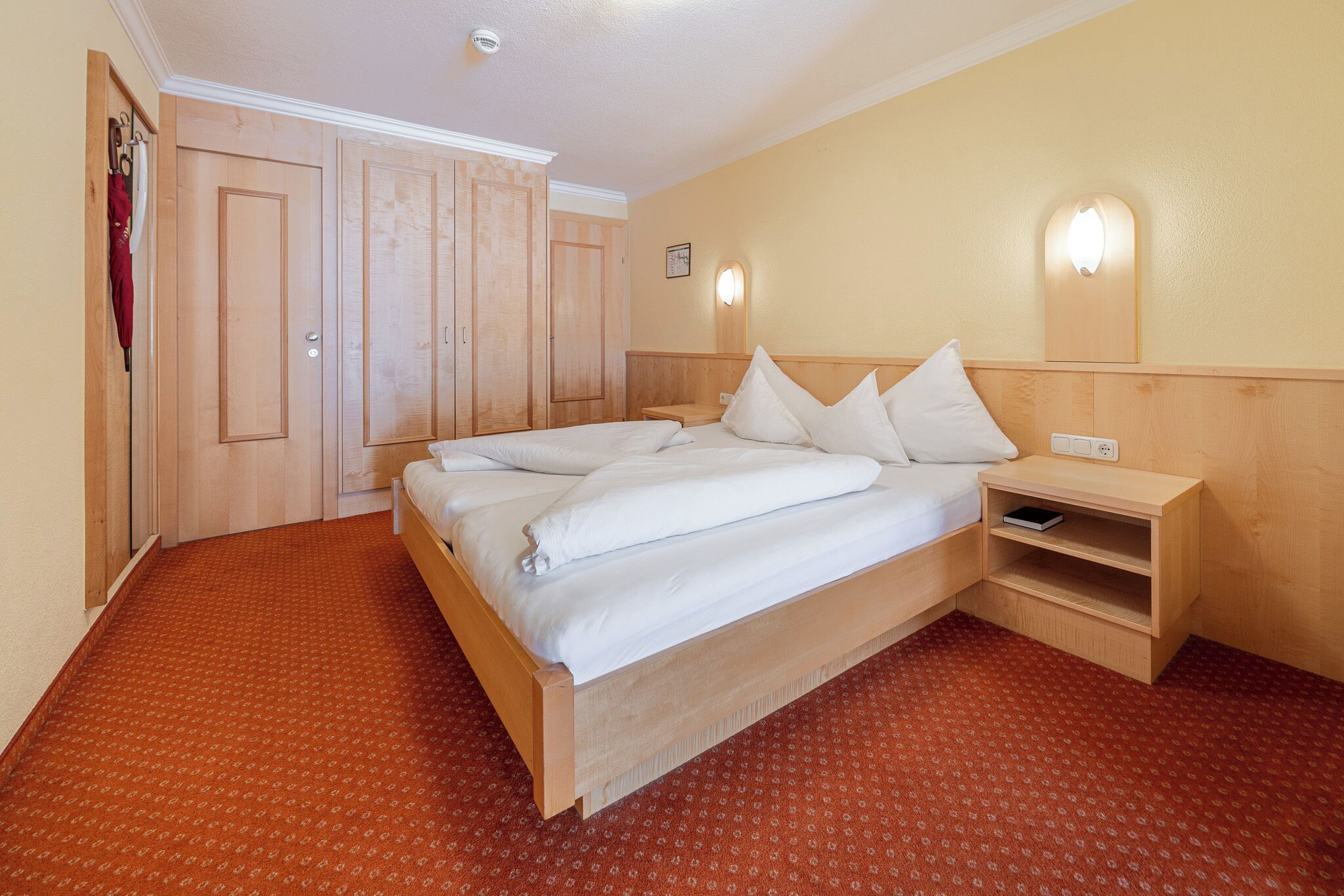 A hotel room with a bed, a nightstand, a wardrobe, and a wall-mounted light.