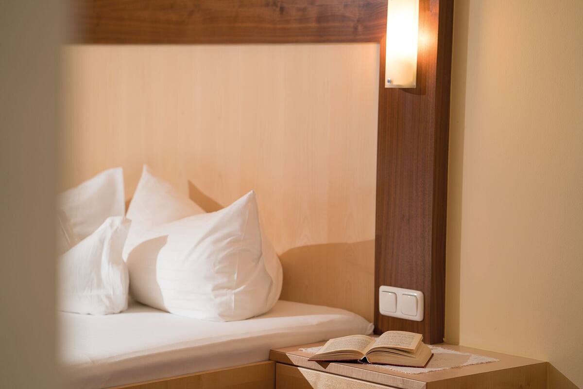 Una cama bien hecha con sábanas y almohadas blancas, una mesita de noche con un libro abierto y una lámpara fijada en la pared.