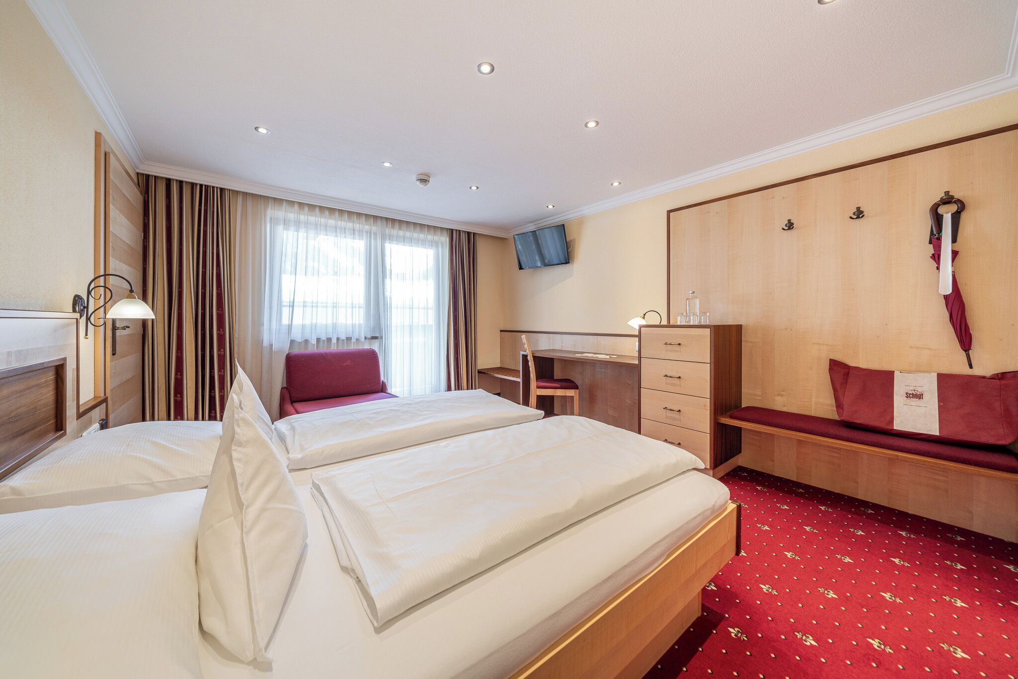 Habitación de hotel con dos camas, un escritorio, una silla, un televisor y una alfombra roja.