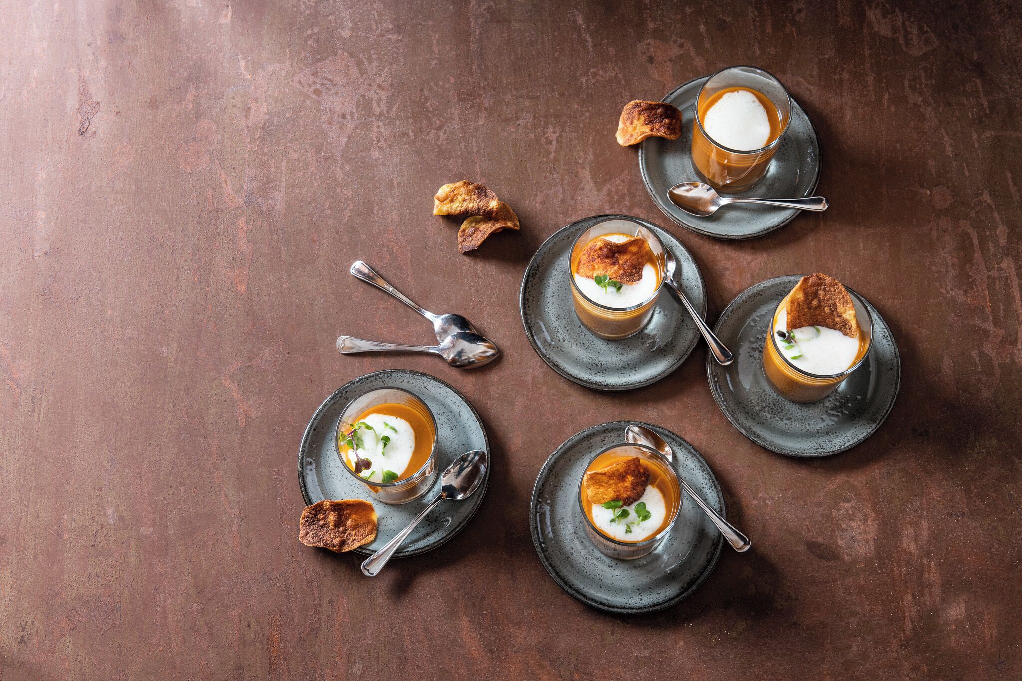 A table with four glasses of soup, each with a different topping, and two spoons on the table.