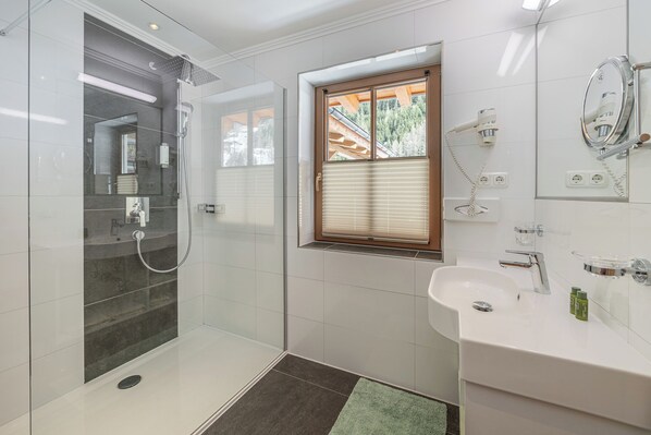A modern bathroom with a glass-enclosed shower, a window with blinds, a sink with a mirror, and a towel rack. - Hotel Schöpf (Laengenfeld)