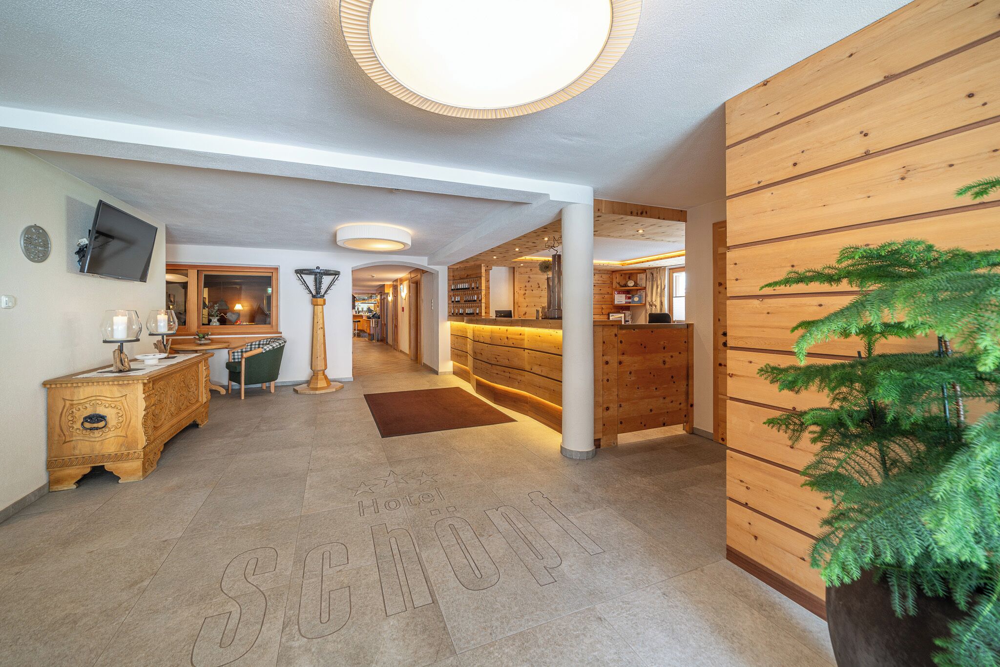 A hotel lobby with a reception desk, a TV mounted on the wall, a floor lamp, and a potted plant.