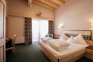 Quarto de hotel com cama de madeira, escrivaninha, cadeira, radiador e janela com cortinas.