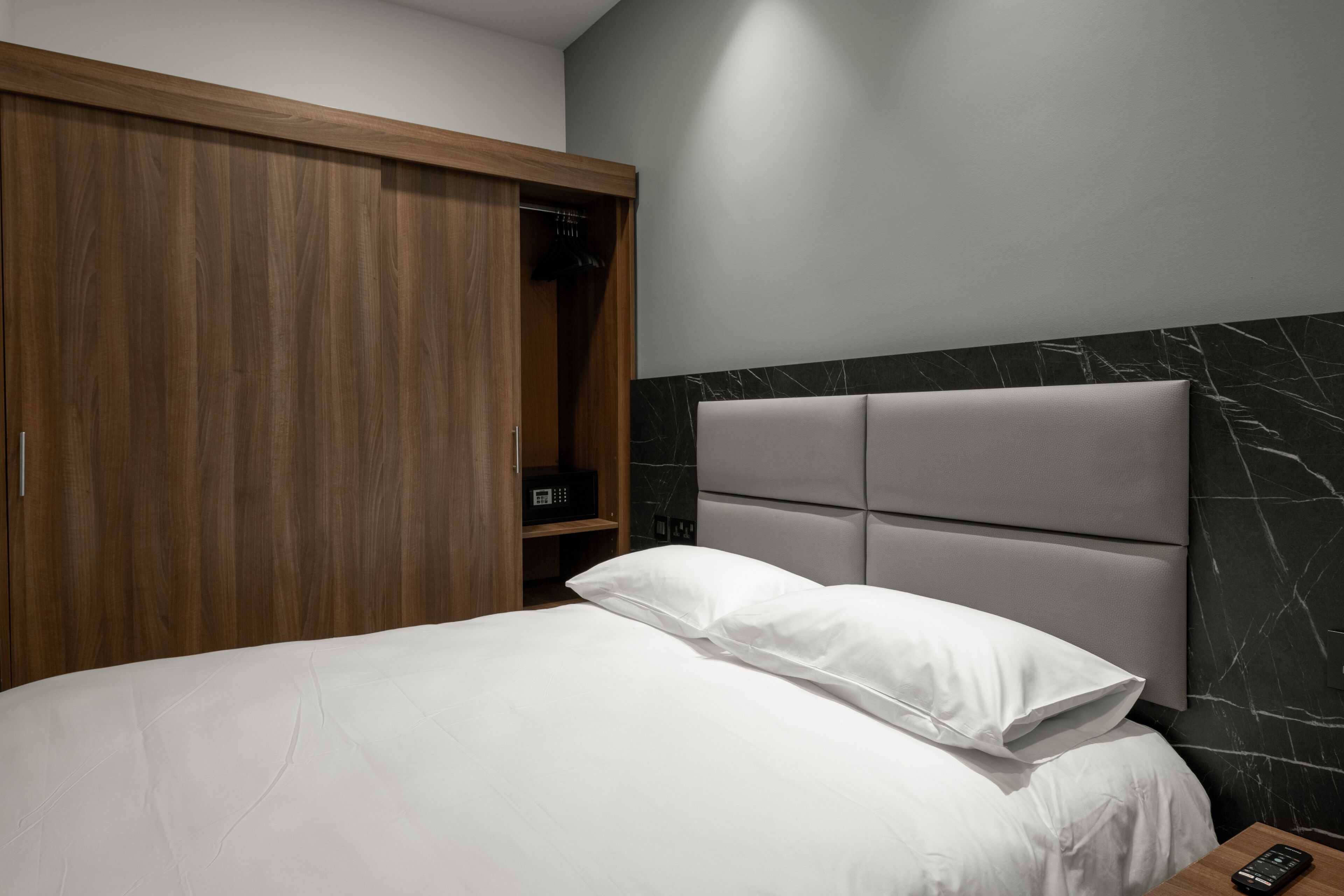 A bedroom with a bed, a wooden wardrobe, a marble headboard, and a bedside table with a remote control.