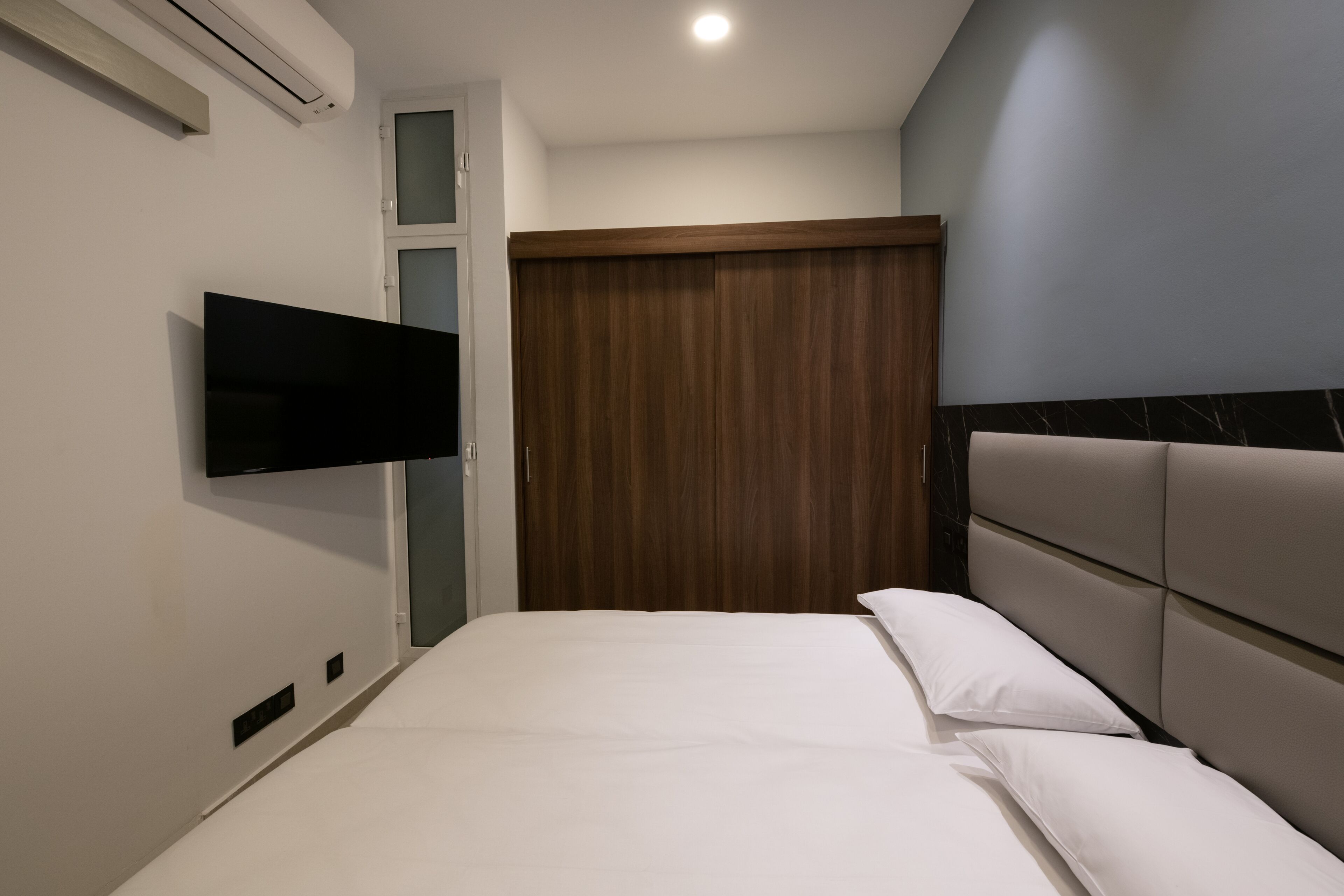 A modern hotel room with a bed, a flat-screen TV mounted on the wall, and a wooden wardrobe.