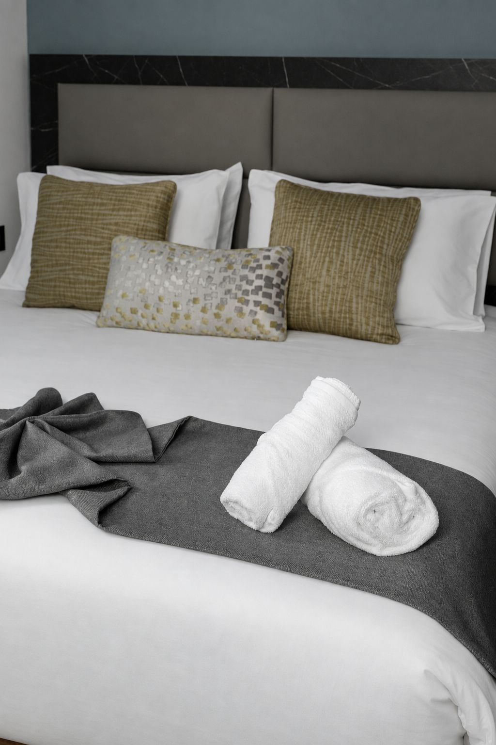 Una cama bien hecha con un edredón gris, dos toallas blancas enrolladas y una almohada estampada.