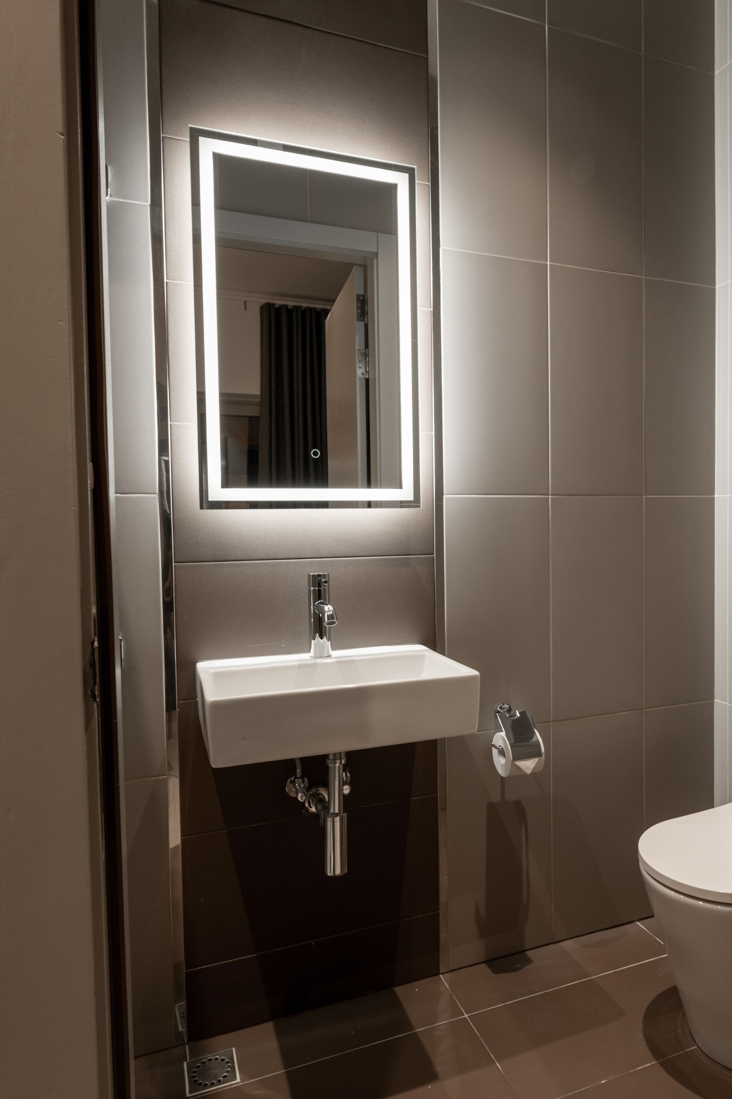 Un baño moderno con un lavamanos blanco, un espejo con iluminación integrada y un inodoro.