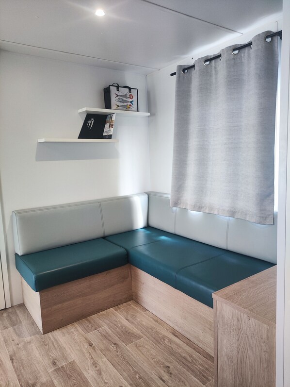 Una habitación moderna y compacta con un banco tapizado en color verde azulado, suelo de madera, una pared blanca con una estantería que sostiene un bolso y un cuadro enmarcado, y una cortina gris.