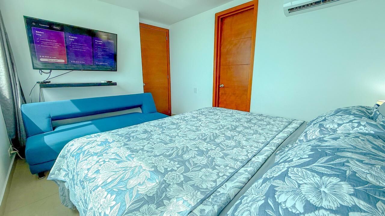 Une chambre à coucher avec un lit, un canapé bleu, un téléviseur fixé au mur et deux portes en bois.