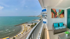 Een balkon met uitzicht op het strand en de zee, een bank met kussens, en een schilderij aan de muur.