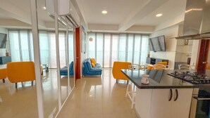 Een moderne keuken met een centraal eiland, een eetgedeelte met oranje stoelen, en een woongedeelte met een blauwe bank en gele kussens.