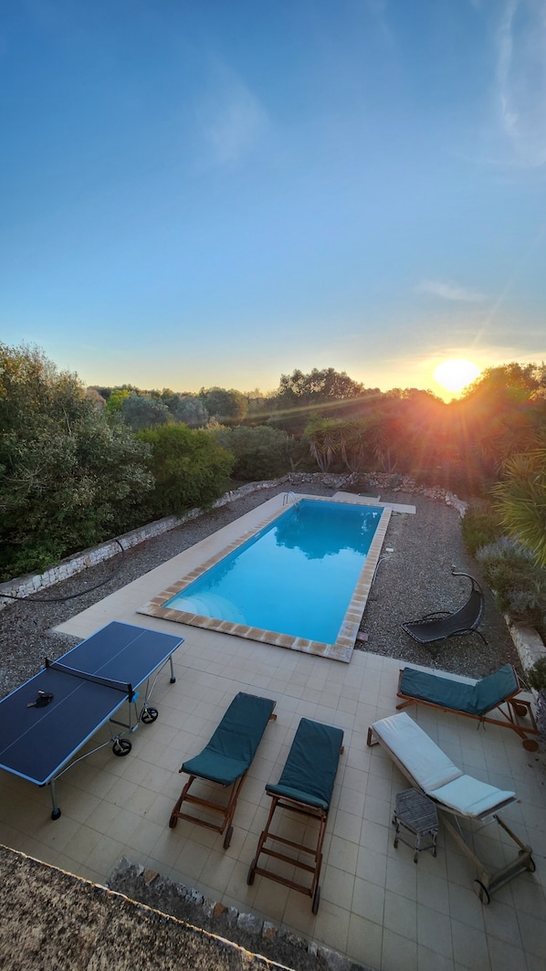 A rectangular swimming pool with a tiled deck, surrounded by greenery and a stone wall. There are lounge chairs and a table with a table tennis table.