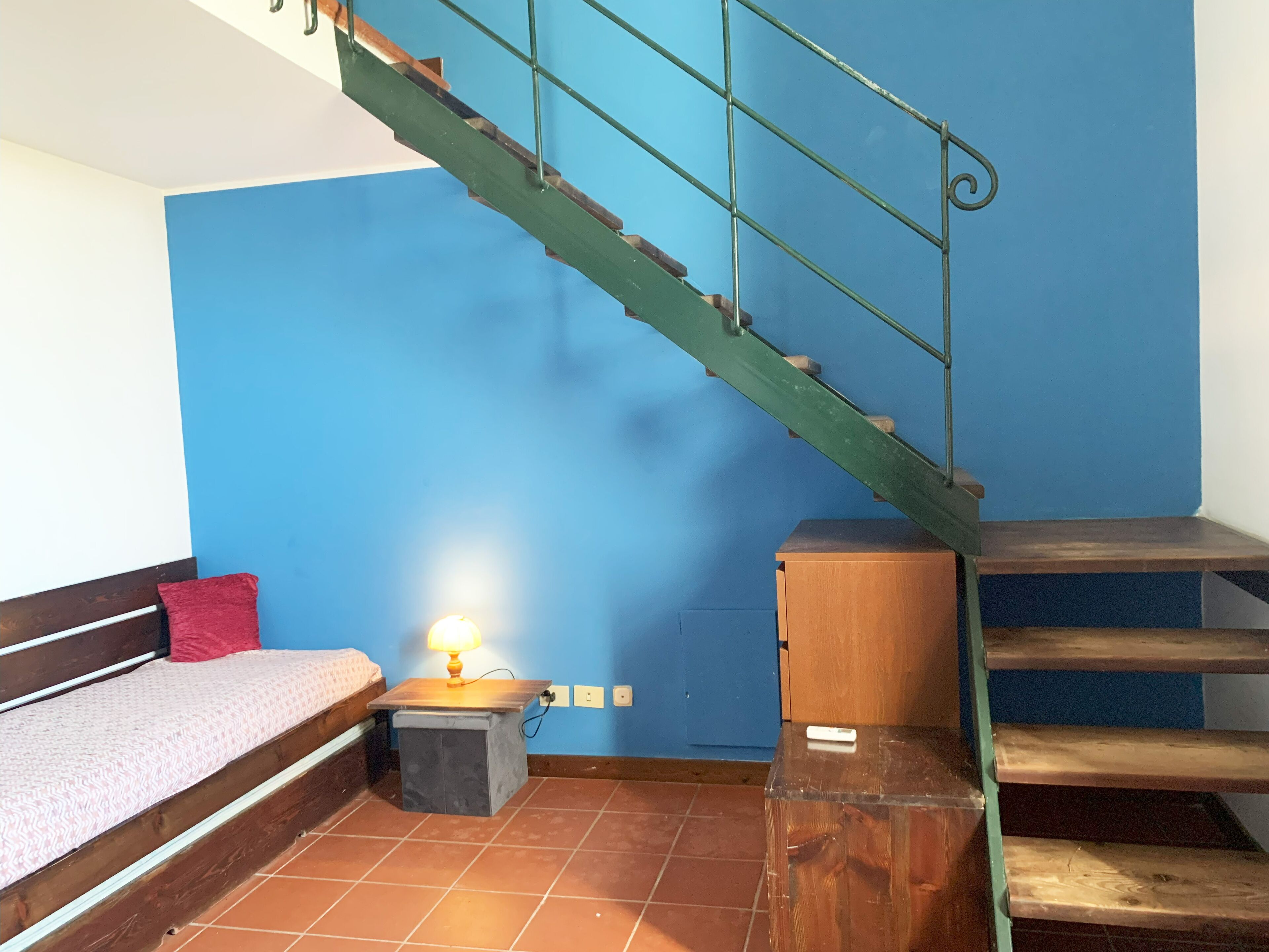 A room with a wooden bench, a small table with a lamp, a staircase, and a blue wall.