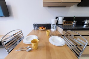 Eine kleine Küche mit einem Holztisch, zwei Stühlen, einer Kaffeemaschine, einer Fruchtschale und einer Wasserflasche.