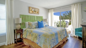 Un dormitorio con cabecera verde, cama grande con ropa de cama estampada, mesita de noche con lámpara y vistas al exterior a través de una puerta corrediza de vidrio.