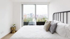Ein Schlafzimmer mit großem Fenster, einem Bett mit weißer Bettwäsche und Zierkissen, einem kleinen Nachttisch mit Pflanze und Blick auf eine Stadtansicht.
