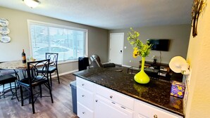 A compact kitchen with a dining area, a large window with blinds, a bright yellow vase with flowers, and a flat-screen TV mounted on the wall. - 2 Bedroom Single Story Townhouse (Beaverton)