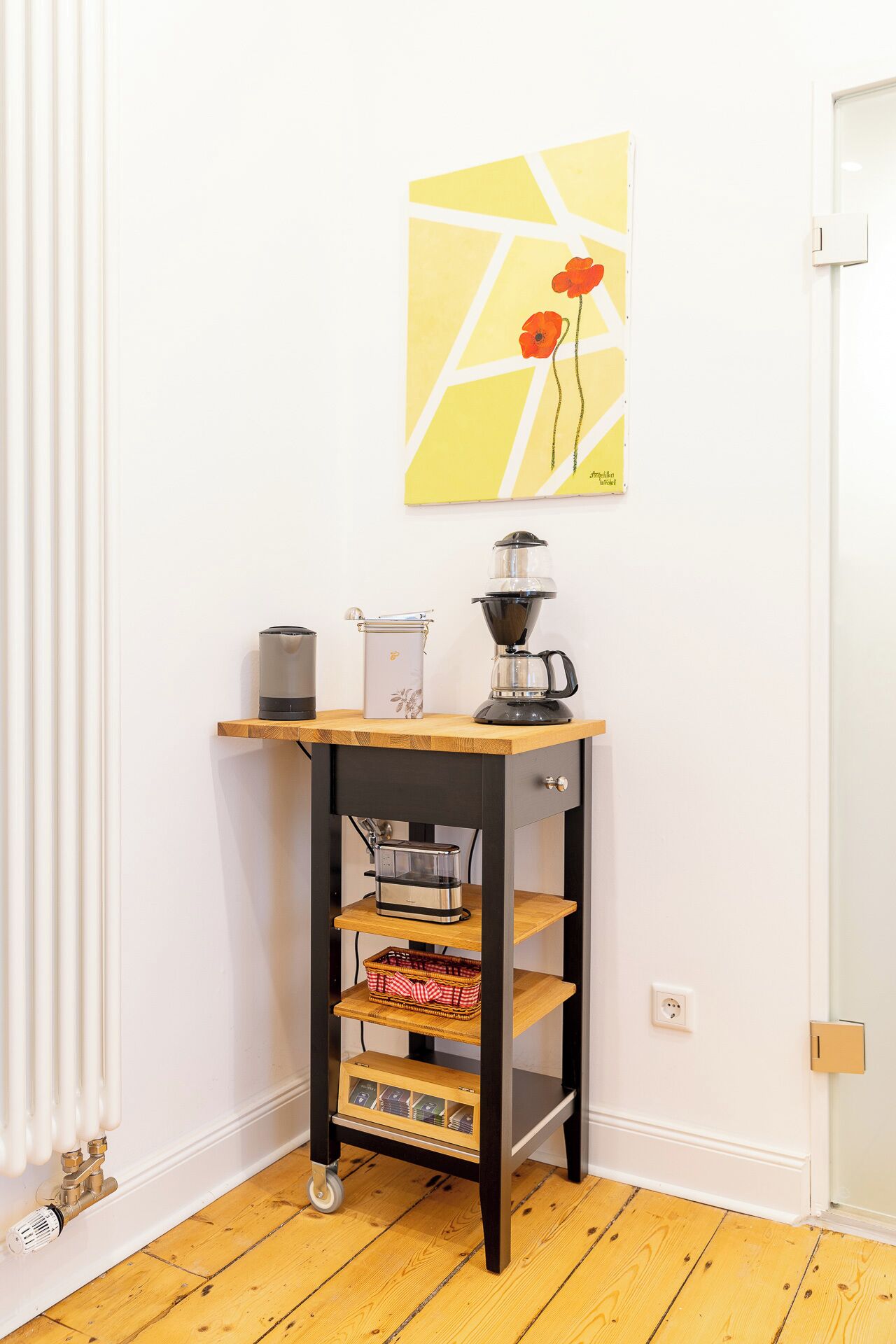 Ein Schreibtisch im Eckbereich mit Kaffeemaschine, einem Korb und einer kleinen Ablage für Bücher.