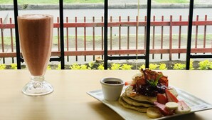 A breakfast setting with a glass of a brown beverage, pancakes with fruit and a small cup of syrup, and a view of a red fence and greenery outside. - Ecohotel Portillo de San Bernardo  (Vergara)