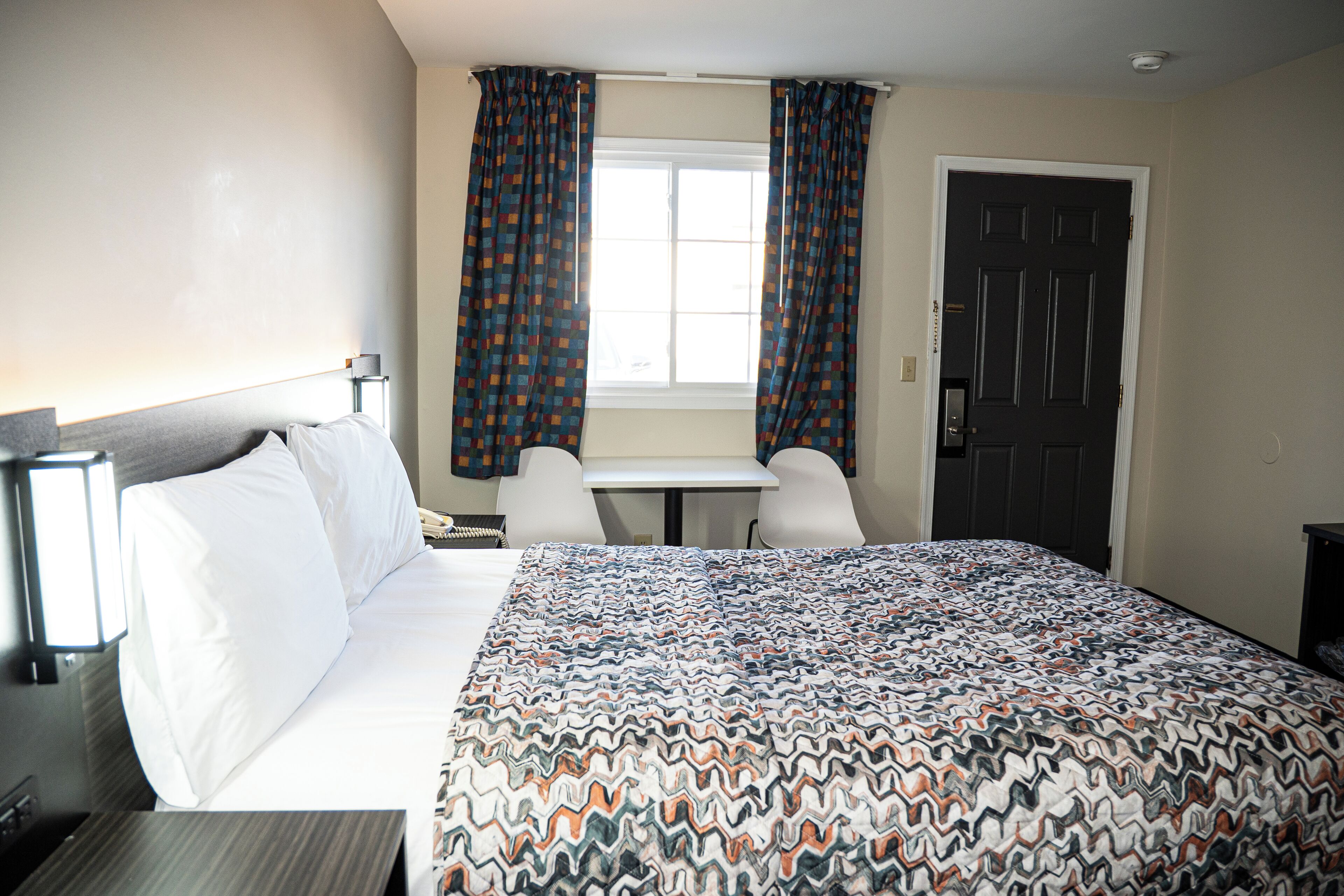 Quarto de hotel com uma cama, uma escrivaninha, duas cadeiras, uma janela com cortinas e uma porta.
