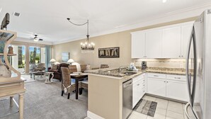 A spacious kitchen with white cabinets, a stainless steel refrigerator, and a dining area with a table and chairs. - Peninsula Links B102 (Gulf Shores)