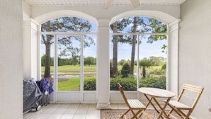 A covered patio with a view of a manicured lawn and trees, featuring a table and chairs. - Peninsula Links B102 (Gulf Shores)
