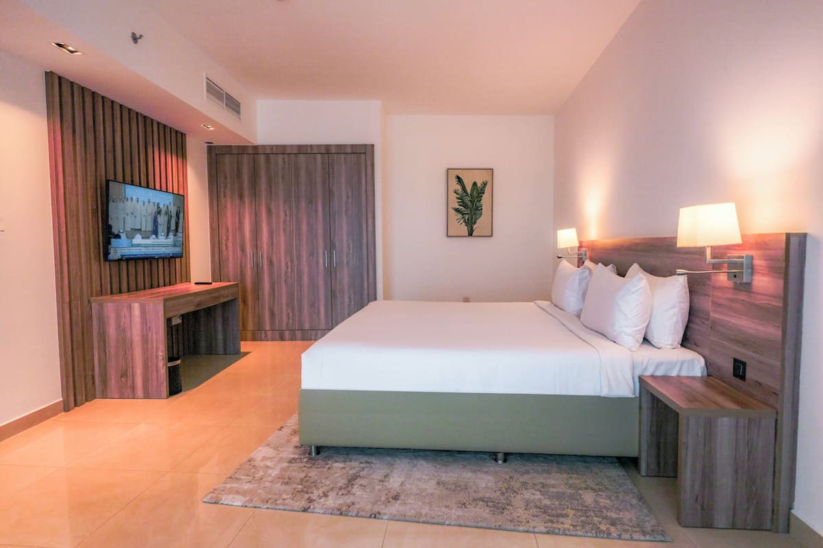 Een moderne hotelkamer met een groot bed, een aan een houten paneel bevestigde televisie, een nachtkastje met een lamp, en een klein bankje.