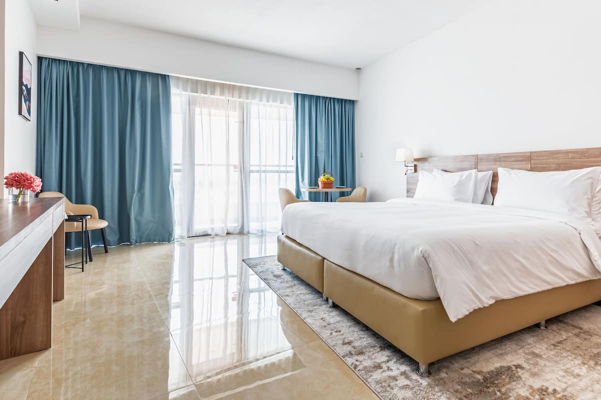 Een hotelkamer met een groot bed, een bureau met stoel, een tafeltje met een mand, en uitzicht op de natuur door grote ramen met blauwe gordijnen.