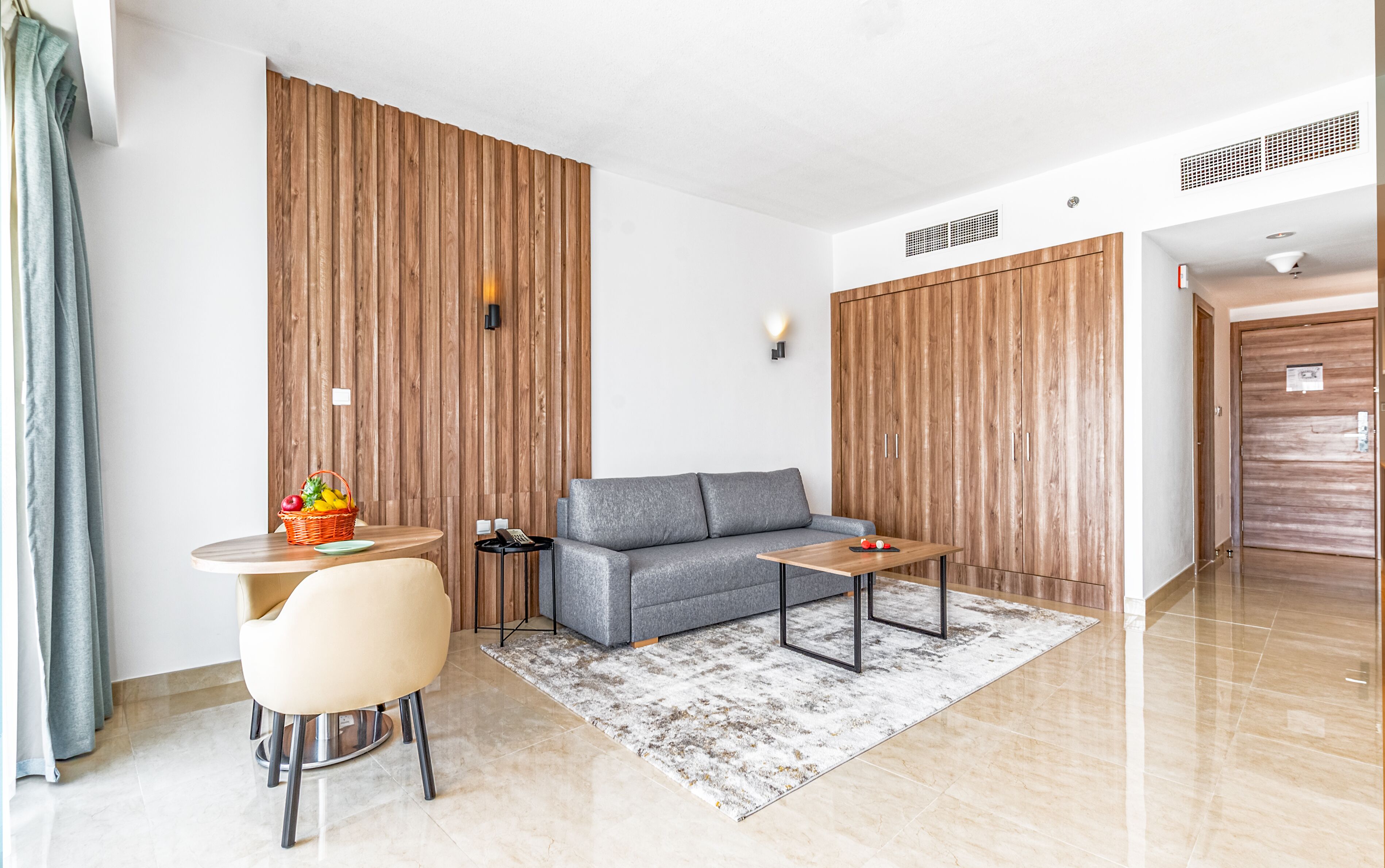 Une chambre d’hôtel moderne avec un canapé gris, une petite table ronde, un fauteuil beige, des murs recouverts de panneaux en bois et une grande armoire.