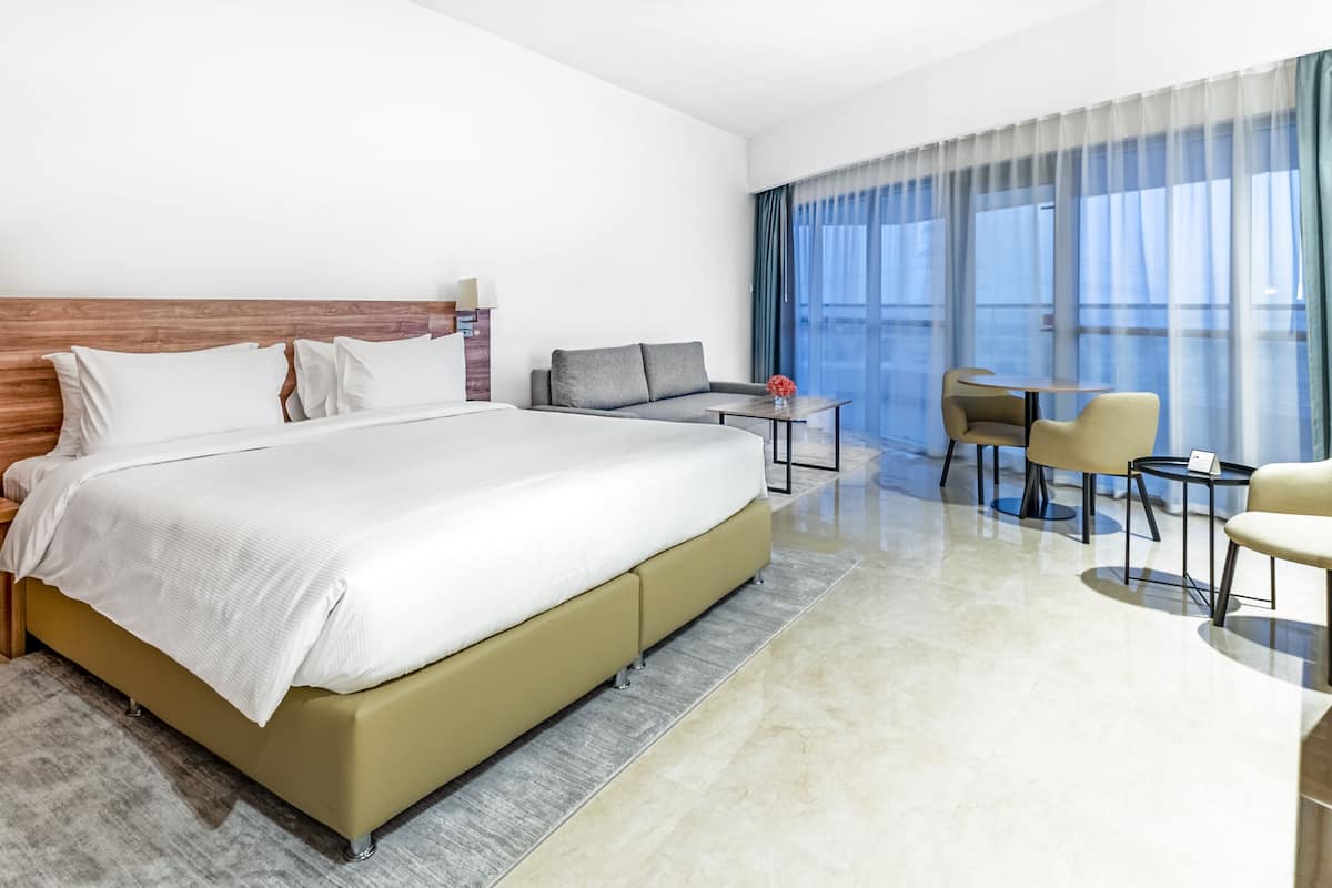 Een moderne hotelkamer met een groot bed, een bureau, een bank en uitzicht op de stad door grote ramen.