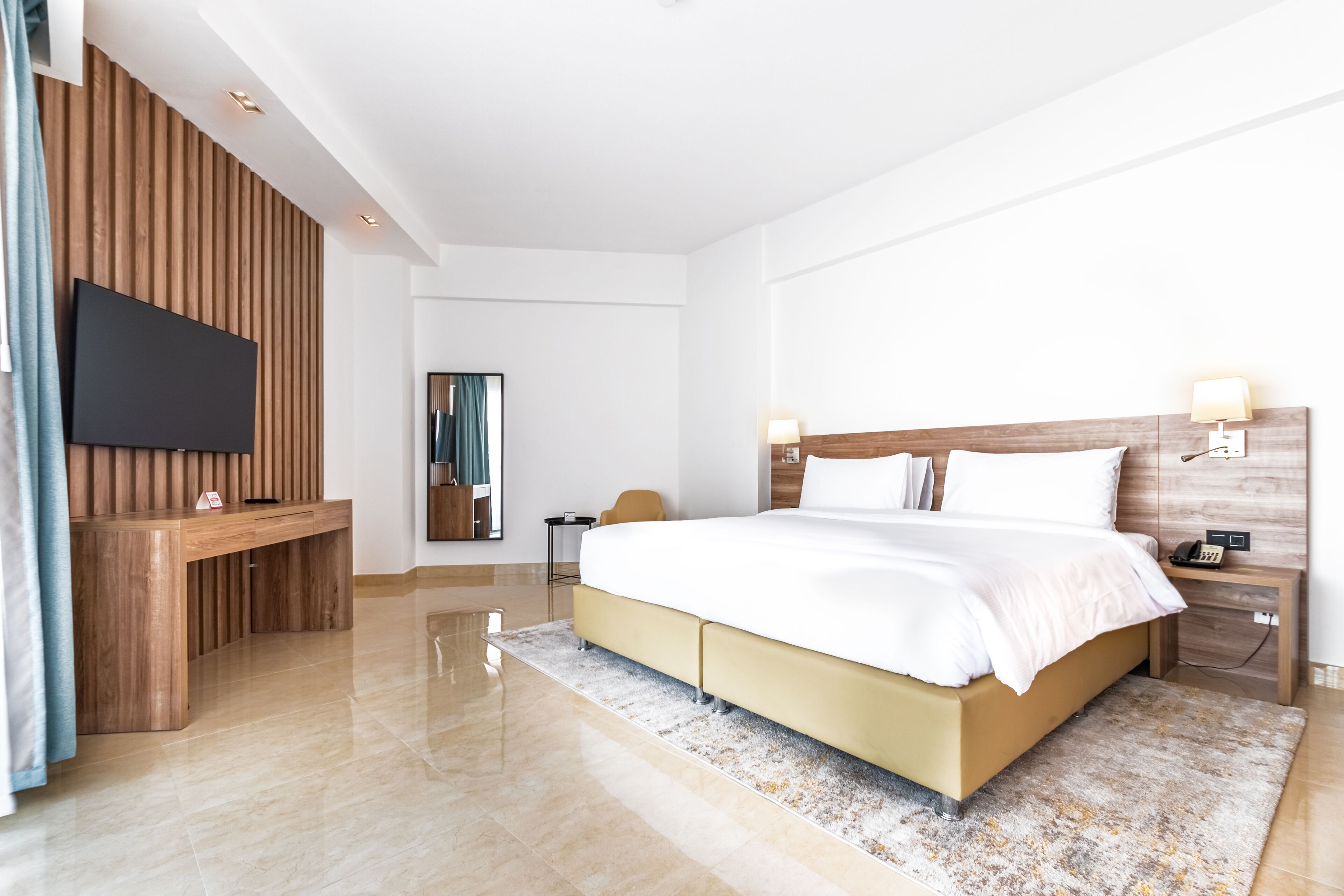 A modern hotel room with a large bed, a flat-screen TV, a wooden desk, and a nightstand with a lamp.