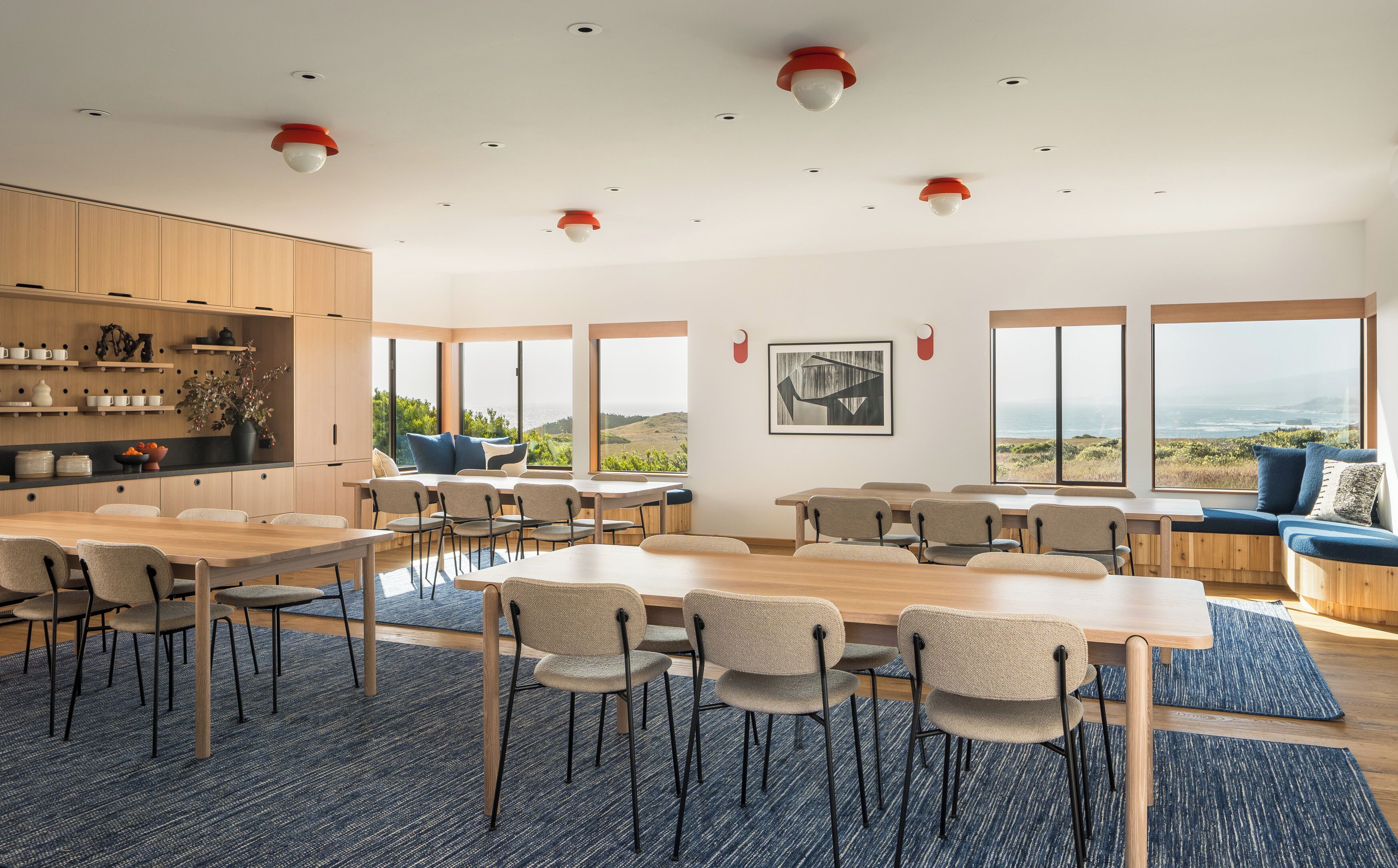 Een ruime eethoek met meerdere tafels en stoelen, grote ramen met uitzicht op de oceaan, en een keuken met houten kasten en planken.