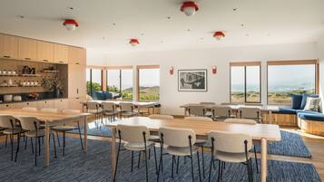 A spacious dining area with multiple tables and chairs, large windows offering ocean views, and a kitchen area with wooden cabinets and shelves.