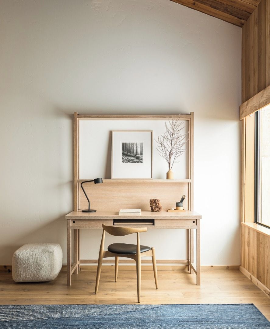 A minimalist workspace with a wooden desk, a chair, a framed picture, and a vase with branches.