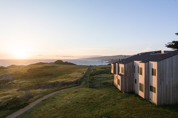 A modern building with a wooden exterior, a pathway leading to the ocean, and a view of the sea and hills in the background.