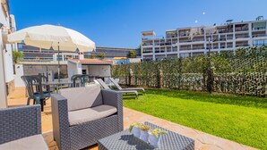 Eine Terrassenfläche mit Korbmöbeln, einem Tisch mit einer Blumenplatte und Blick auf ein mehrgeschossiges Gebäude im Hintergrund.