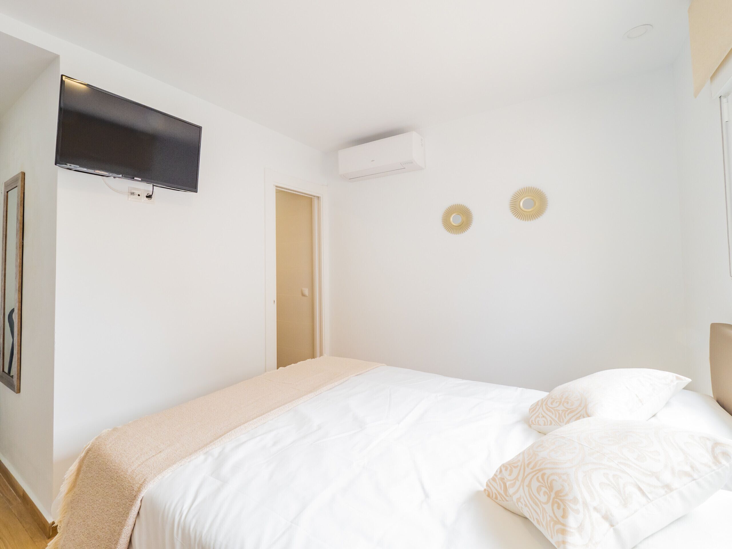 A bedroom with a bed, a mounted TV, an air conditioning unit, and two wall-mounted decorative lights.