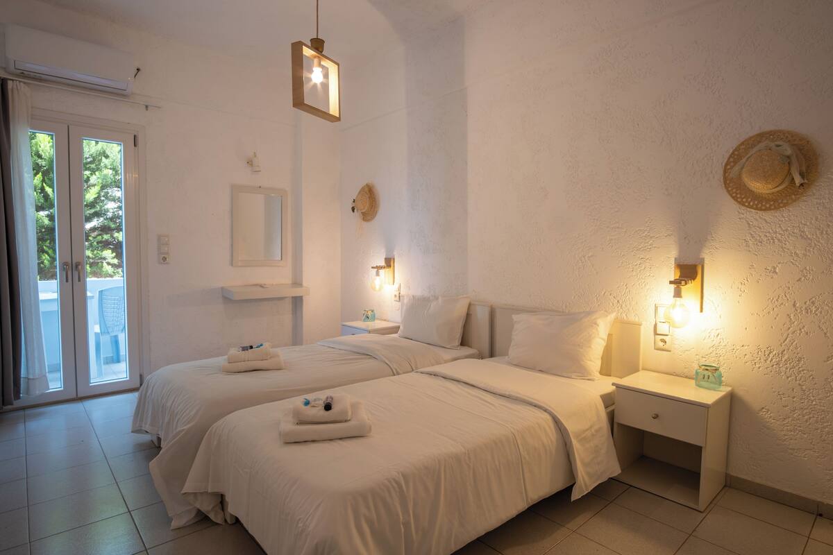 A hotel room with two beds, a nightstand, a wall-mounted hat, and a sliding glass door leading to an outdoor area.