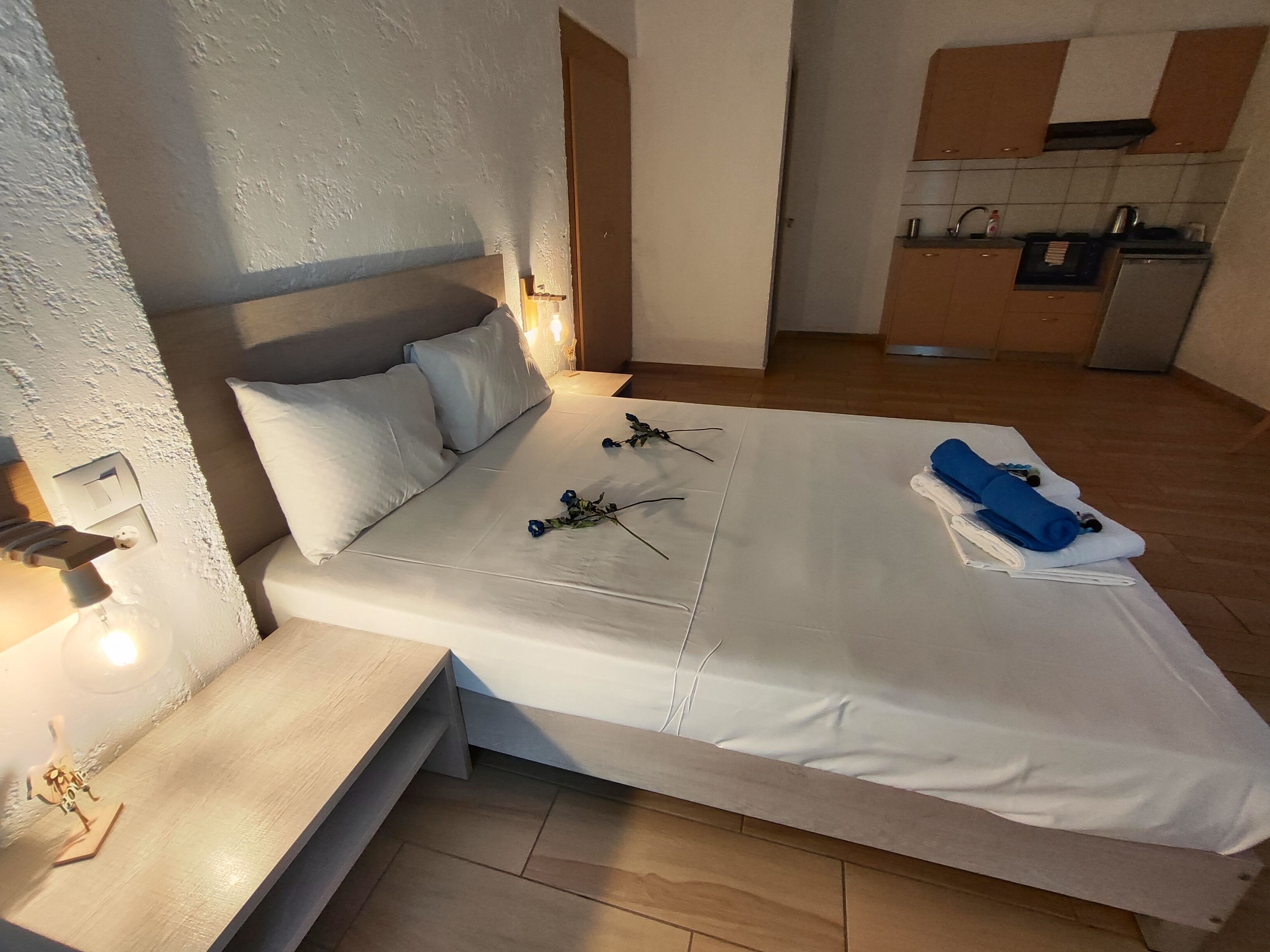 A bedroom with a bed, bedside table, and a small desk. There is a kitchenette in the background.
