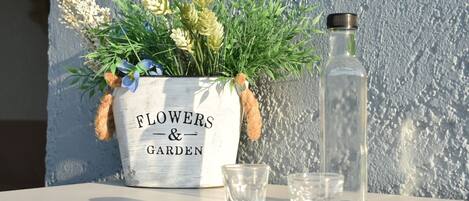A white planter with 'Flowers & Garden' text, containing green plants and blue flowers, next to a clear bottle and three glasses on a table against a textured wall.