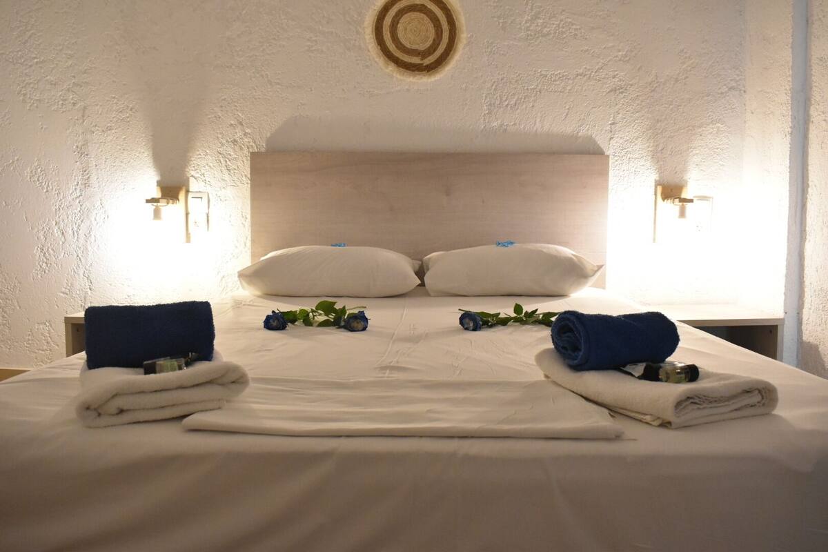 A neatly made bed with white linens, two rolled towels, and a decorative blue rose.