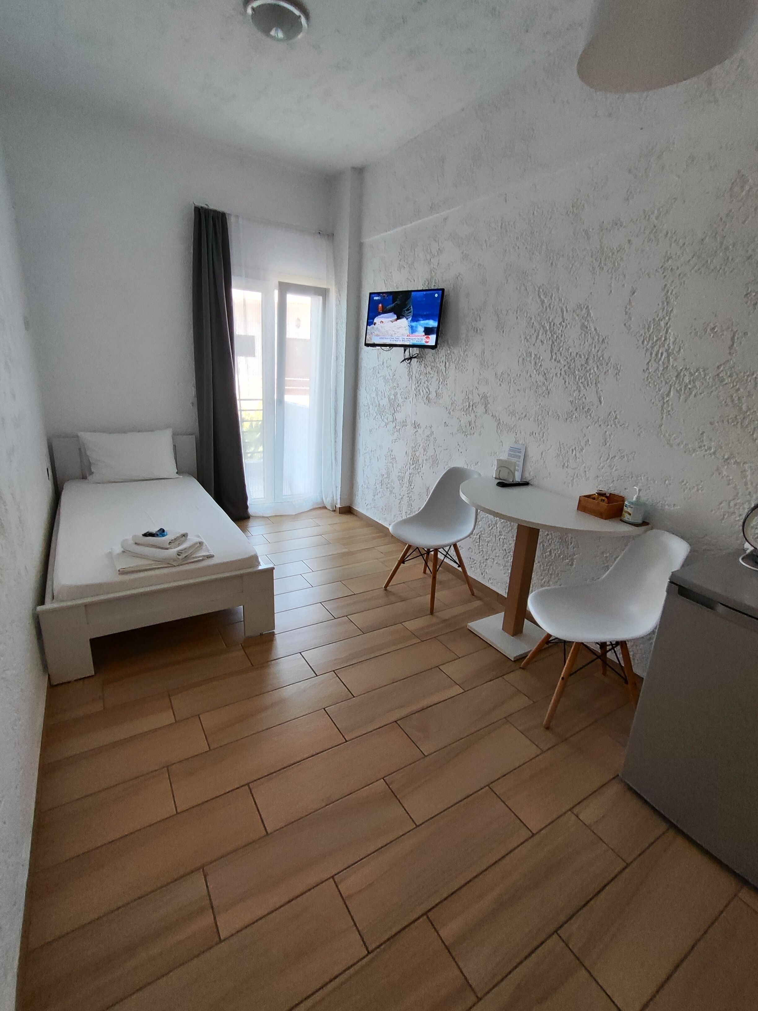 A modern hotel room with a bed, a desk, two chairs, a television, and a small kitchenette.