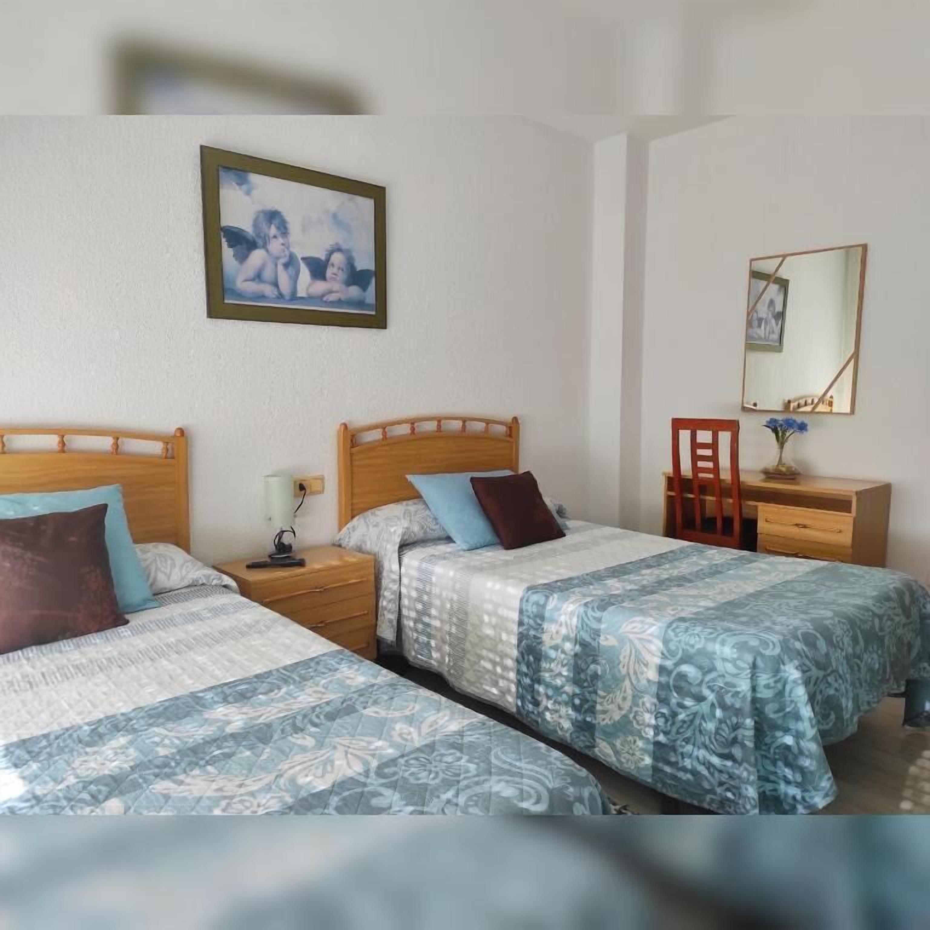 A bedroom with two single beds, a nightstand with a lamp, a chair, a desk with a mirror, and a framed picture on the wall.