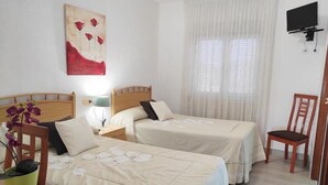 Een hotelkamer met twee bedden, een aan de muur bevestigde televisie, een raam met dunne gordijnen en een potplant op het nachtkastje.
